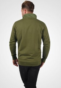 Olivgrüner Sweatshirt mit hohen Kragen, gerippten Bündchen und Saum, aus einem weichen Stoff gefertigt. Getragen mit dunklen Jeans, von hinten betrachtet.