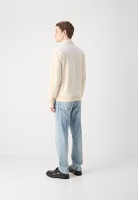 Beige fleecetröja med en kontrasterande vit krage, kombinerad med ljusblå jeans och svarta snörskor; sedd från baksidan.