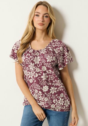 Kvinde med langt blondt hår iført en lilla blomstret kortærmet bluse og blå jeans, stående op ad en hvid væg.