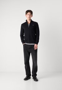 Svart hettegenser med glidelås og høy krage, brukt over en stripete skjorte. Kombinert med mørke jeans og svarte sko. Enkel design, ingen synlige mønstre.