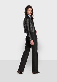 Black leather cropped jacket with zippered pockets, paired with black trousers. Model stands sideways against a neutral background.
