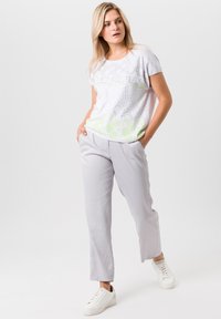 Witte t-shirt met een groen en grijs cirkelpatroon, gecombineerd met lichtgrijze broek. Model draagt witte sneakers. Casual fit, korte mouwen.