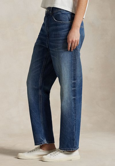 Persona que lleva vaqueros azules de pierna recta y zapatillas blancas, de pie frente a un fondo neutro con una camisa blanca ligeramente visible.