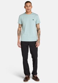 Ljusblå t-shirt med kort ärm, rund halsringning och liten logotyp på vänster bröst, ihop med svarta byxor och bruna skor.