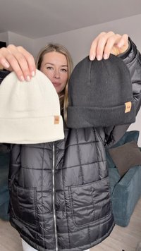 Dvě čepice jsou drženy nahoře: jedna krémová, jedna černá. Obě jsou pletené s ribovanou texturou, mají ohrnutý lem a malou hnědou nálepku vpředu.