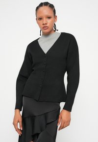 Černý kardigan s V-výstřihem a dlouhými rukávy, který má zapínání na knoflíky. Tkanina vypadá měkce a má hladkou strukturu.