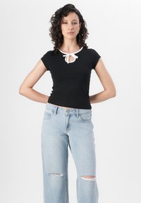 Juoda trumpomis rankovėmis palaidinė su rakto skylutės iškirpte ir balta juostele, derinama su šviesiai mėlynomis suplėšytomis platėjančiomis džinsomis. Lygus fonas.