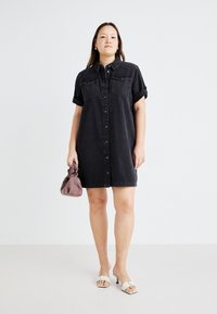 Zwart denim jurk met korte opgerolde mouwen, knoopsluiting aan de voorkant, borstzakken en een losse pasvorm. Gecombineerd met een paarse tas en witte sandalen.