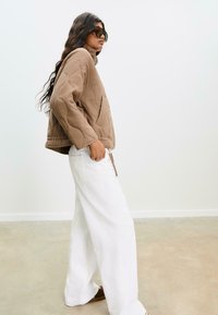 Veste matelassée marron avec un col montant et deux poches avant, associée à un pantalon large blanc ample et des sandales marron.