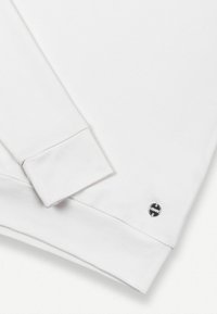 Povečava manšete in roba bele dolgo rokavne jopa z majhnim okroglim kovinskim črno-belim logotipom blagovne znamke blizu roba.