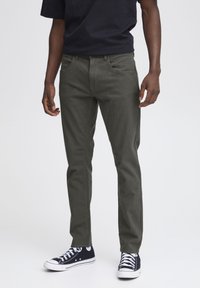 Pantalones ajustados de color verde oliva oscuro con una textura suave, cuentan con bolsillos estándar y costuras blancas, combinados con zapatillas negras de caña alta.