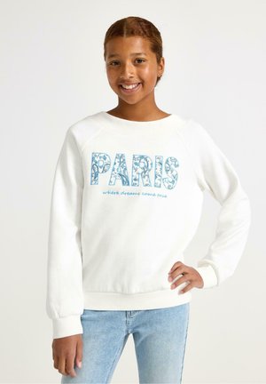 Lächendes Mädchen mit geflochtenen Haaren, das einen weißen Pullover mit der Aufschrift "PARIS, wo Träume wahr werden" und hellblaue Jeans trägt, steht mit einer Hand in der Hüfte.