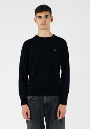 Maglione nero a maglia con scollo rotondo e maniche lunghe. Presenta un piccolo logo sul petto. Indossato con jeans in denim grigio scuro.