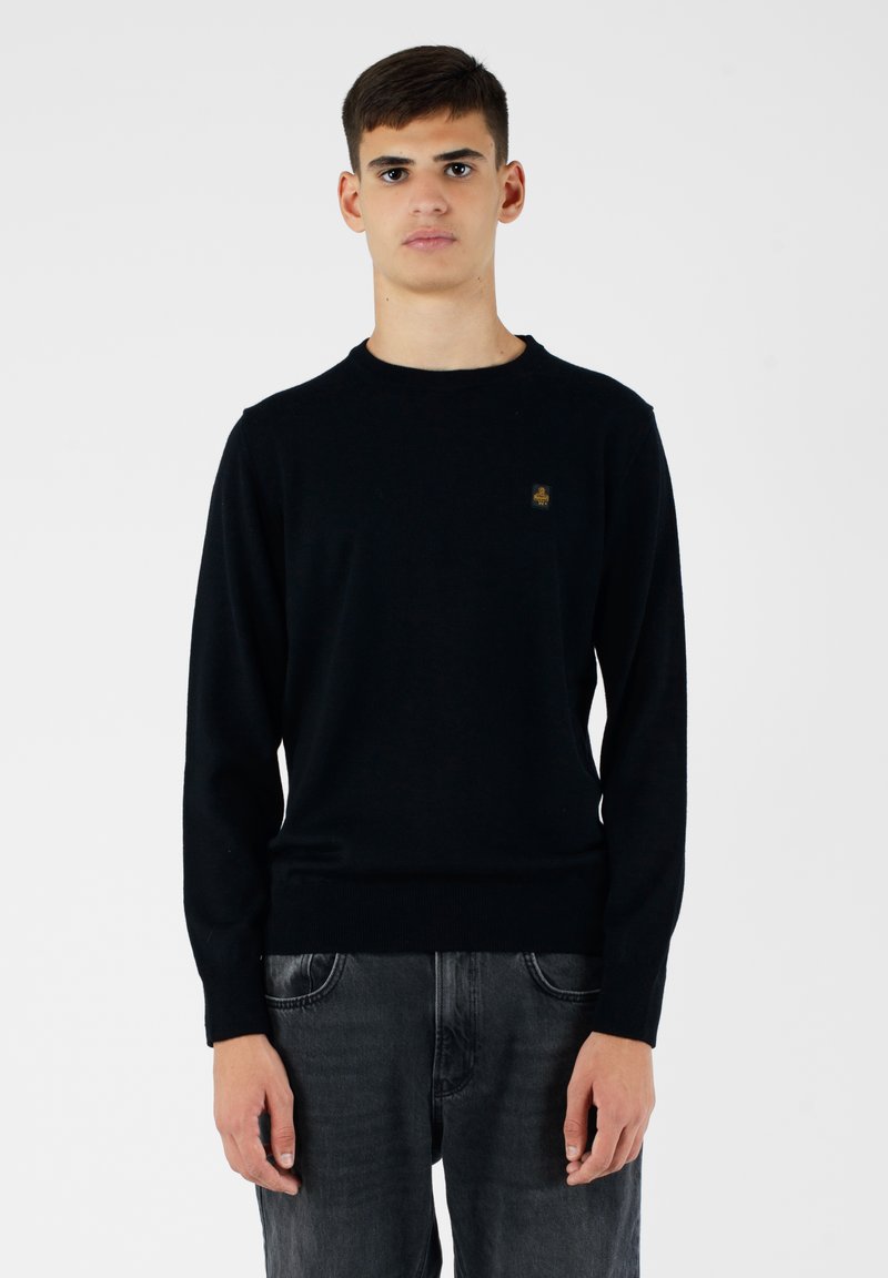 Maglione nero a maglia con scollo rotondo e maniche lunghe. Presenta un piccolo logo sul petto. Indossato con jeans in denim grigio scuro.