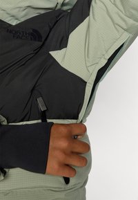 Šviesiai žalia izoliacinė striukė su juodu interjeru. Yra tinklelio raštas, šoninis kišenė su užtrauktuku ir The North Face logotipas.