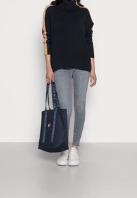Sac cabas bleu marine avec des anses à texte, arborant un petit logo. Porté avec un col roulé noir et un jean skinny gris, associé à des baskets blanches.
