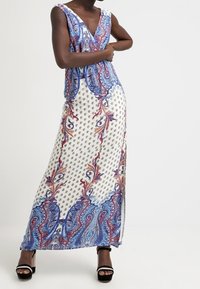 Robe maxi avec décolleté en V, motif cachemire bleu et rouge sur fond blanc, dotée de fentes latérales et d'un tissu léger. Sandales à talons noires.