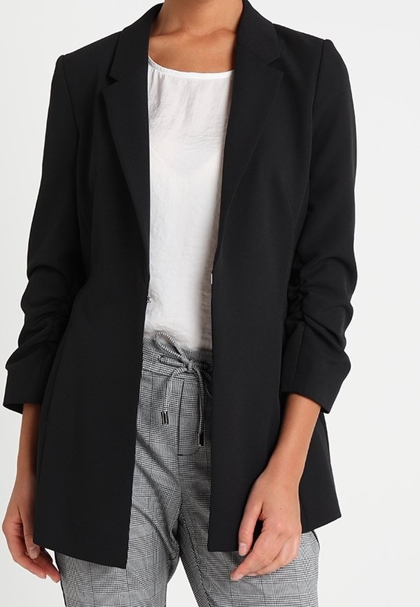 Schwarzer Blazer mit tailliertem Design, langen Ärmeln und offenem Vorderteil. Getragen über einem weißen Oberteil und karierter Hose. Glatte Textur.
