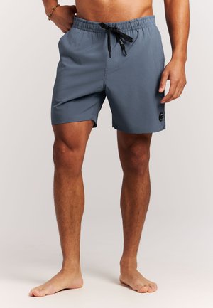 Man draagt blauw zwemshorts tot halverwege de dij met een zwarte trekkoord, staat blootsvoets op een effen lichte achtergrond.