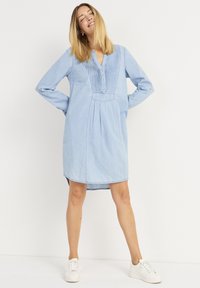 Lichtblauwe chambray jurk met lange mouwen, knoopsluiting, geplooide details en een ontspannen pasvorm, gecombineerd met witte sneakers.