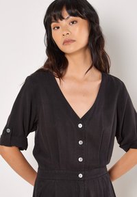 Mono negro con botones en la parte delantera y escote en V profundo, mangas cortas enrolladas y cinco botones contrastantes. Tejido suave y texturizado.