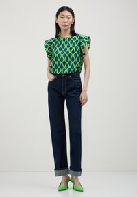 Donna che indossa un top verde a fantasia con maniche a volant, jeans scuri a risvolto e scarpe verdi con tacco alto, in piedi davanti a uno sfondo neutro.