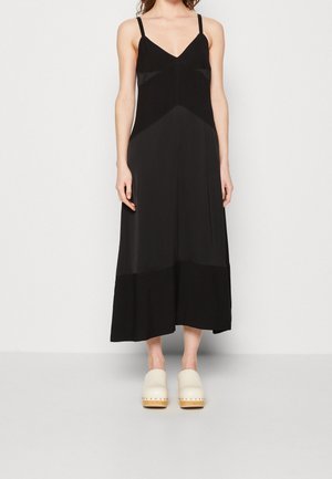 Femme portant une robe midi noire sans manches avec un décolleté en V et des sabots blancs sans lacets, debout devant un fond clair uni.