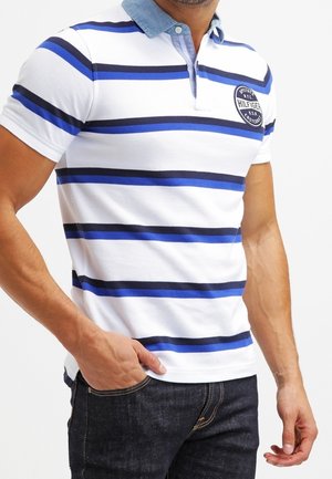 Kortærmet polo shirt i hvid med blå og marineblå vandrette striber. Har en denimkrave og logo-patch på venstre bryst.
