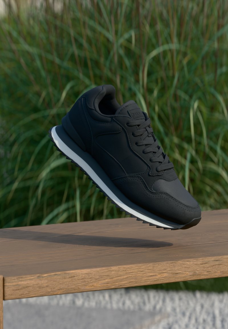 Musta urheilukenka, jossa on tekstuurilla varustettu päällinen, nauhakiinnitys ja erivärinen valkoinen välipohja; esillä ilmassa puupinnan yllä.