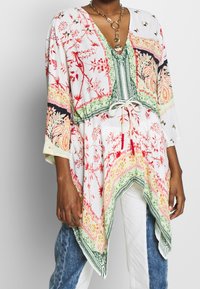 Žena v tunice s květinovým vzorem v bílé, zelené a červené barvě, s elastickým pasem a asymetrickým lemem, doplněná dvoubarevnými džíny a bílými kalhotami.