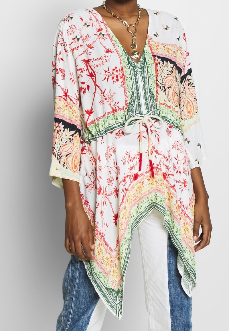 Žena v tunice s květinovým vzorem v bílé, zelené a červené barvě, s elastickým pasem a asymetrickým lemem, doplněná dvoubarevnými džíny a bílými kalhotami.