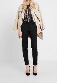 Trench-coat beige porté sur un haut bleu marine à motifs avec chaînes dorées, associé à un pantalon noir fuselé et des chaussures à talons noires.