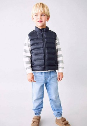 Laikku kashmir valkoinen puffer-liivi, joka on käytetty raidallisen harmaan ja valkoisen pitkähihaisen paidan päällä, yhdistettynä vaaleansinisiin käännettyihin farkkuihin ja ruskeisiin kenkiin.