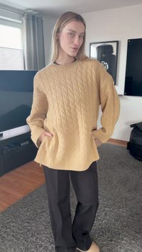 Άτομο που στέκεται μέσα σε κλειστό χώρο, φορώντας μπεζ πουλόβερ με πλεκτό σχέδιο καλώδιο και μαύρο παντελόνι, με τα χέρια στις τσέπες του πουλόβερ, ουδέτερη έκφραση.