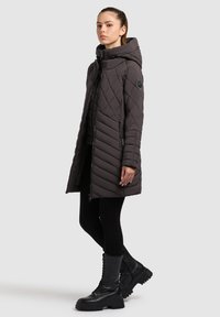 Young woman wearing a quilted dark gray hooded winter coat, black leggings, and black lace-up combat boots standing at slight angle.