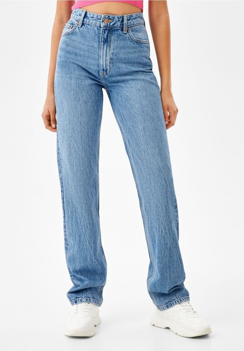 Femme portant un jean bleu clair taille haute, coupe droite, des baskets blanches et un top court rose, debout devant un fond blanc.