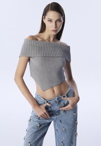 Šedý žebrovaný crop top s odhalenými rameny a přiléhavým střihem, doplněný o světle modré džíny se stříbrnými ozdobami.