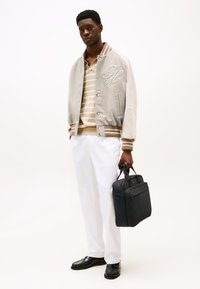 Veste bomber gris clair avec des accents crème et un logo brodé, polo rayé beige et blanc, pantalons blancs et un sac noir. Chaussures noires.