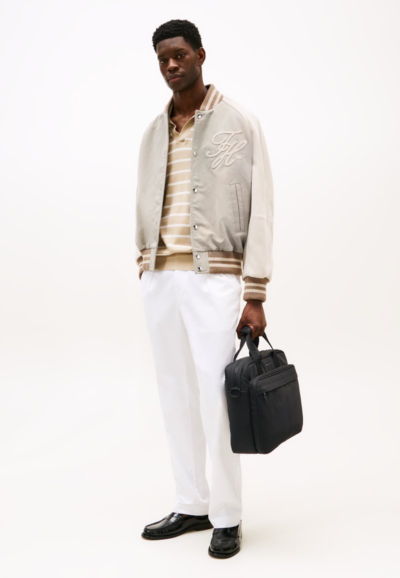 Veste bomber gris clair avec des accents crème et un logo brodé, polo rayé beige et blanc, pantalons blancs et un sac noir. Chaussures noires.