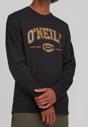Persoon met een zwart shirt met lange mouwen waarop in goud de tekst "O'NEILL Santa Cruz California" op de borst staat, armen gekruist ter hoogte van de taille.
