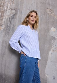 Camicia a righe azzurre e bianche con colletto arricciato, maniche lunghe e polsini elastici, abbinata a jeans di denim blu a vita alta.