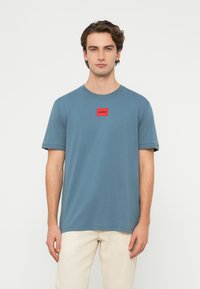 Kék pamut póló környakú, rövid ujjú, mellrészen piros négyzet alakú „HUGO” logós címkével. Egyszerű dizájn és kényelmes szabás.