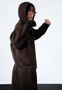 Sweatshirt à capuche marron avec une poche avant, des poignets côtelés et une coupe décontractée, associé à un pantalon assorti avec des poches latérales.