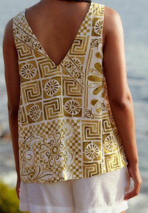 Person wearing a sleeveless top with yellow and white geometric and floral patterns, paired with white shorts, standing near water.