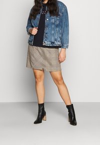 Mujer con chaqueta de denim azul, top negro, pantalones cortos de cuadros y botas de tacón negro hasta el tobillo, de pie contra un fondo claro y liso.