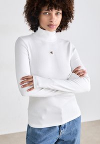 Witte geribbelde lange mouw top met een hoge kraag en een klein logo detail op de borst. Zachte textuur en getailleerd ontwerp.