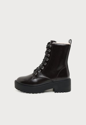 Schwarzer Leder-Schnürstiefel mit klobiger Sohle und Zuglasche, seitlich gezeigt vor hellgrauem Hintergrund.