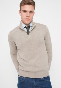 Beżowy sweter z pół-zamkiem, ściągaczami przy rękawach, o fakturowanym splocie, zestawiony z jasnoniebieską koszulą i wzorzystym krawatem.