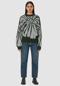 Maglione a maglia di colore verde scuro con un grande motivo floreale grigio, maniche lunghe e vestibilità ampia. Abbinato a jeans blu e stivali neri.