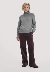 Frau steht in einem grauen Rollkragensweater, dunkelvioletten Cordhosen und dunkelbraunen Schuhen vor einem schlichten hellen Hintergrund.